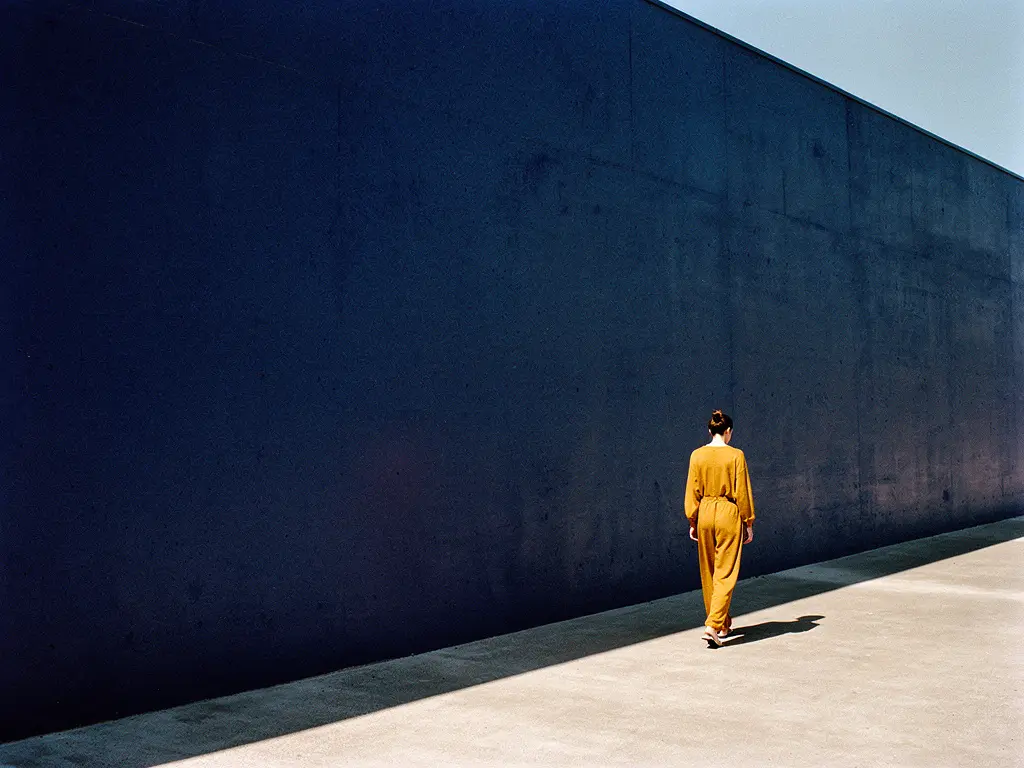 A person walking past a long deep-navy concrete facade in hard sunlight, seen from behind.
