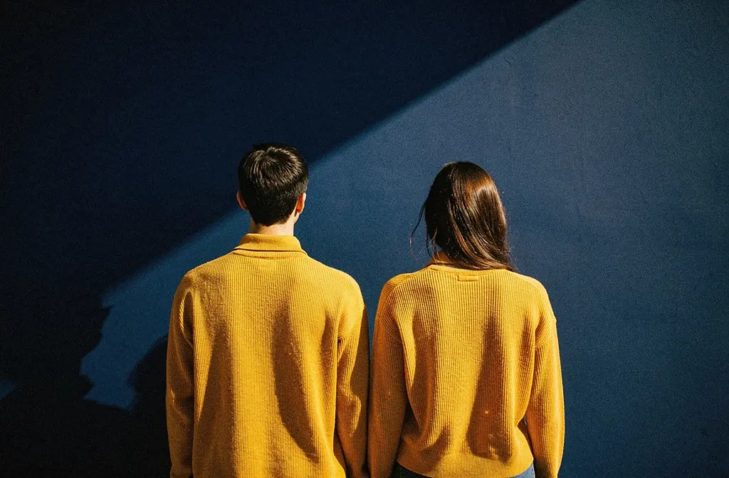 Two people photographed from behind in amber knitwear against a deep navy wall, a hard diagonal shadow across it.